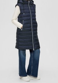 Lange, marineblaue Steppweste mit Kapuze, Reißverschluss vorne und Seitentaschen, kombiniert mit weit geschnittenen Jeans und weißen Plateausneakern.