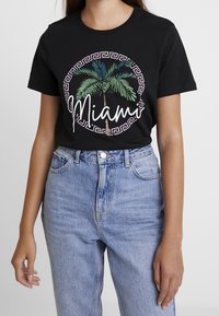 Sort bomulds T-shirt med en rund grafik, der har palmer og teksten "Miami" i hvid og grøn, bæres sammen med lyseblå denimjeans.