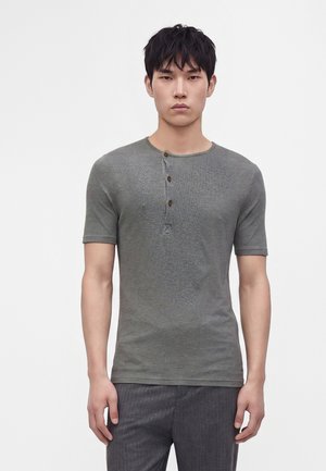 Jeune homme aux cheveux courts noirs portant un t-shirt henley gris à manches courtes et un pantalon gris foncé sur un fond blanc uni.