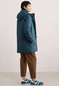 Teal waterdichte parka met capuchon, zijzakken en trekkoord. Gecombineerd met een paar bruine corduroybroek en blauwe suède sneakers met witte strepen.
