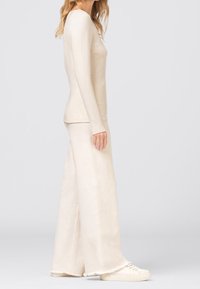 Kremowa bluzka z długim rękawem zestawiona z kremowymi, szerokimi spodniami. Tkanina wydaje się miękka, o gładkiej fakturze z ozdobnymi surowymi brzegami na dole.