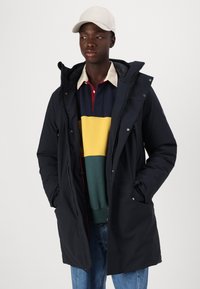Parka blu scuro con cappuccio, dotato di zip frontale, chiusure a pressione e tasche laterali. Indossato sopra un maglione a righe multicolore e jeans blu.