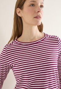 Femme à la peau claire et aux cheveux châtain clair portant un chemisier à rayures longues violettes et blanches, regardant légèrement vers le haut et à droite.