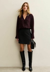 Blusa bordeaux con motivi a cuore neri, abbinata a una mini gonna nera con spacco laterale e stivali alti in pelle nera fino al ginocchio.