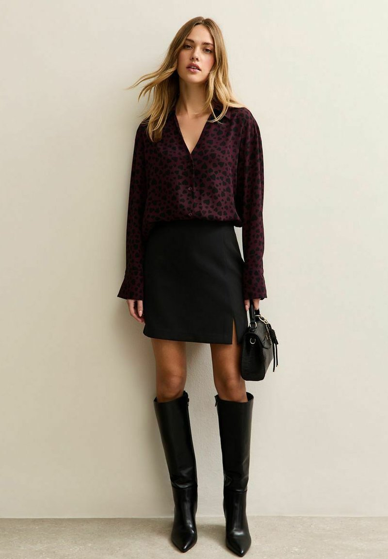 Blusa bordeaux con motivi a cuore neri, abbinata a una mini gonna nera con spacco laterale e stivali alti in pelle nera fino al ginocchio.