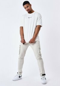 Camiseta de algodón blanca con cuello redondo y pequeño logotipo negro en el pecho, combinada con pantalones cargo beiges claros y zapatillas blancas con detalles negros.