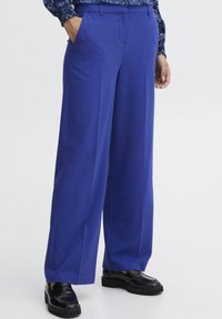 Brede pantalon van felblauwe stof met een gladde textuur, voorzien van zijzakken en een tailored fit, gecombineerd met zwarte instappers.