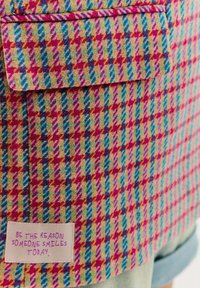 Detailní záběr na barevnou houndstooth tkaninu s kapsou a štítkem s nápisem "Buďte důvodem, proč se někdo dnes usměje."