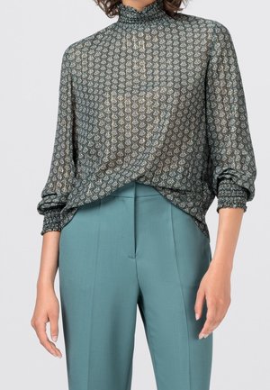 Blouse verte en soie avec un col haut, présentant un motif géométrique en or et bleu. Coupe ample avec des poignets froncés et un détail drapé.