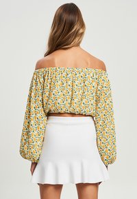 Top corto scollato con motivo floreale giallo, maniche lunghe a sbuffo e dettagli arricciati, abbinato a una mini gonna bianca a corolla.