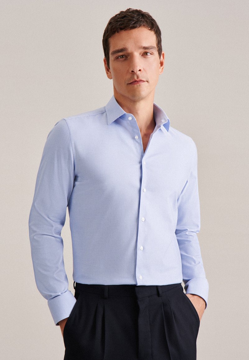 Camicia azzurro chiaro a maniche lunghe con un motivo testurizzato, colletto classico e bottoni bianchi, abbinata a pantaloni scuri.