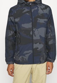 Marinblå jacka med kamouflagemönster, huva, dragkedja och elastiska muddar. Tillverkad av lätt material och har tre ränder på ärmarna.