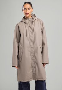 Beige sade takki vettä hylkivällä pinnalla, hupulla, nepparikiinnityksellä, sivutaskuilla ja löysällä leikkauksella. Mallilla esitelty yksinkertaista taustaa vasten.