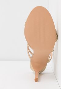 Vue de la semelle d'une sandale beige à talon haut avec un talon fin et un dessous texturé uni sur fond blanc.