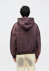 Camisola com capuz em tecido polar de roxo intenso com um desenho desbotado, capuz largo e bainha crua. Apresenta costuras visíveis e acessórios metálicos no capuz.