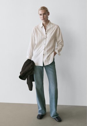 Kobieta ubrana w luźną, białą koszulę w paski na guziki, niebieskie dżinsy i czarne buty, trzymająca ciemną kurtkę w jednej ręce na jednolitym tle.