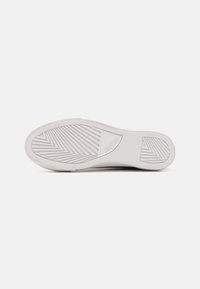 Fehér sneaker talp, texturált, zig-zag mintázattal. Középen a "GUESS" logó dombornyomott. Simább élek és lekerekített forma, minimális részletezéssel.