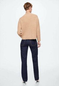 Personne portant un pull beige, un jean bleu foncé et des chaussures blanches à talons hauts, debout de dos sur un fond blanc uni.