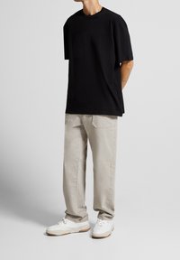 Mies, joka vie mustaa löysää t-paitaa, beigeitä housuja takataskuilla ja valkoisia lenkkareita yksinkertaista taustaa vasten.