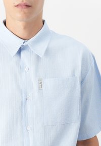 Light blue striped short-sleeve shirt with a pointed collar, front button closure, and a single chest pocket. Features a brand label on the pocket.