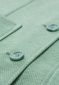Tel fabricado de color verde claro con textura, que presenta dos botones a juego. La vista en primer plano enfatiza el patrón del tejido y los detalles de los botones.