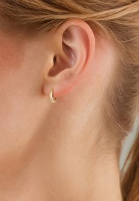 Aukso auskaras žiedo pavidalu, kuris dėvimas ausyje, lygus paviršius, apvalios formos, vidutinio dydžio, su subtiliu blizgesiu neutralios odos tonų fone.