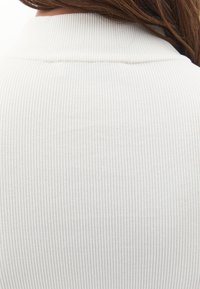 Primer plano de la espalda de una persona que lleva una camisa de manga larga blanca de canalé, con el cabello castaño liso visible en la parte superior.