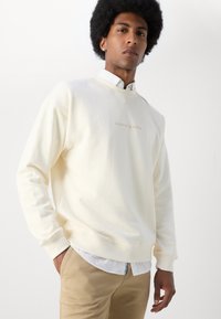 Cremefarvet sweatshirt lavet af blødt stof, med rund halsudskæring og broderet logo. Sammen med en hvid skjorte med krave nedenunder.