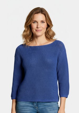 Kvinde med skulderlangt, bølget lysebrunt hår iført en blå striktrøje og blå jeans, som smiler let mod en ensfarvet hvid baggrund.