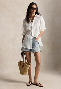 Nő, aki túlméretezett fehér, gombos ingben, farmer rövidnadrágban, fekete szandálokban, napszemüvegben, vörös nyaklánccal, nagy fonott bevásárlótáskával a vállán.