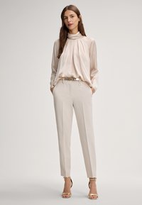 Blusa plissettata di un leggero beige con colletto alto; abbinata a pantaloni cremosi su misura e sandali con tacco dorato. Leggera lucentezza su tutta la superficie.