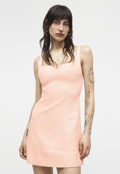 Femeie cu păr lung și întunecat și tatuaje, purtând o rochie roz deschis texturată fără mâneci, stând în fața unui fundal alb simplu.