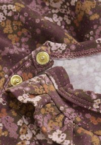 Lila blomsterprydd tyg med volangkant och gyllene tryckknappar, med en blandning av rosa, gult och grönt. Mjuk textur.