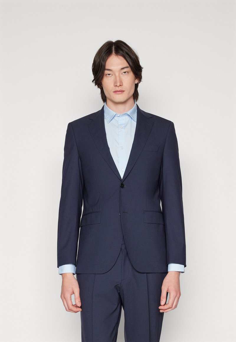 Marineblauer Anzug mit taillierter Passform, strukturierten Revers, einem Knopf und zwei Taschen. Hellblaues Hemd darunter mit einem spitzen Kragen.