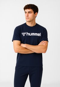 Młody mężczyzna w granatowej odzieży sportowej marki Hummel, stoi z założonymi rękami, patrząc w bok na jednolitym białym tle.