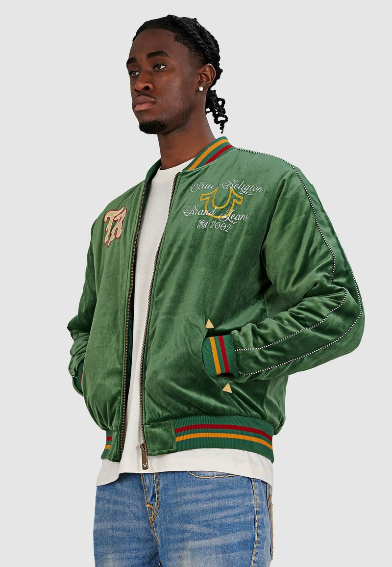 Groene fluweel bomberjack met geborduurde logo's, gestreepte geribde manchetten en zoom, en metalen studs langs de mouwen. Ritsclosure.