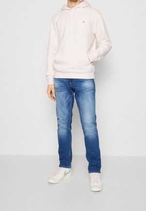 Mies, joka pukeutunut hennon vaaleanpunaiseen huppariin, haalistuneisiin sinisiin farkkuihin ja valkoisiin lenkkareihin, joissa on vaaleanvihreä Nike-logo ja vaaleanpunaiset nauhat, seisoo yksinkertaisen taustan edessä.