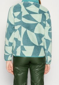 Chaqueta de forro polar en tonos verde menta claro y azul verdoso con patrón geométrico abstracto; cuello redondeado, mangas largas y dobladillo elástico. Combinada con pantalones verdes.