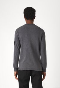 Graues Langarmshirt mit strukturiertem Strickdesign, Rundhalsausschnitt, gerippten Bündchen und einem Patch am linken Ärmel. Leicht abgerundeter Saum.