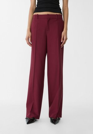 Femme portant un pantalon bordeaux à taille haute et jambes larges, associé à des chaussures noires à bouts pointus et un haut noir sans manches.