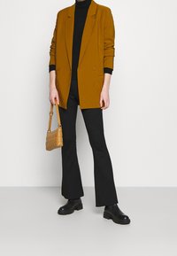 Personne portant un blazer moutarde, un col roulé noir, un pantalon noir évasé, des bottes noires, tenant un petit sac matelassé couleur tan sur un fond blanc.