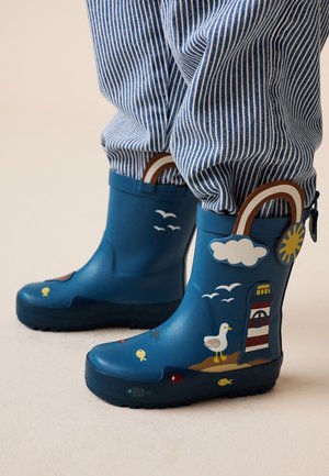 Bambino che indossa stivali da pioggia blu con motivi di faro, uccelli marini, pesci, sole e nuvole, abbinati a pantaloni a righe blu e bianche.