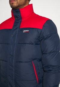 Man som bär en vadderad jacka från Tommy Jeans i marinblått och rött med hög krage och tryckknappar, som visar bröst- och axelområdet.