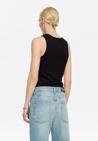 Črna brezrokavnik na osebi, v kombinaciji s svetlo modrimi jeans hlačami. Top ima klasičen kroj z okroglo ovratnikom in gladko teksturo.
