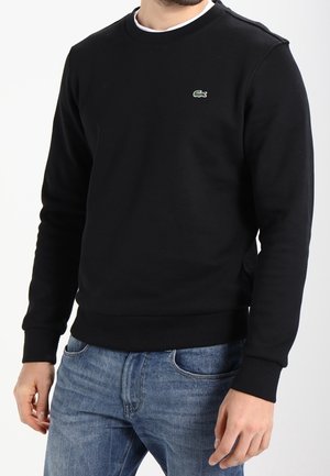 Mann trägt schwarzen Lacoste Langarm-Pullover und blaue Jeans, steht vor einem schlichten weißen Hintergrund.