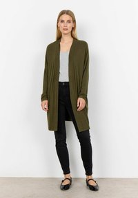 Cardigan lungo verde oliva con apertura frontale, polsini a costine e fessure laterali, indossato sopra una maglietta grigia e jeans skinny neri. Scarpe basse nere.