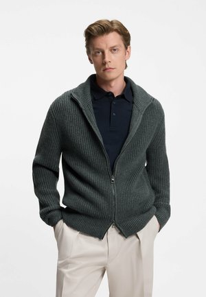 Cardigan a coste grigio scuro con colletto alto e zip frontale, indossato sopra una maglietta polo blu navy, abbinato a pantaloni di colore chiaro.