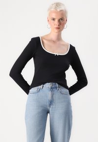 Svart langermet topp med bred utringning, hvit kant og en liten sløyfe i midten av brystet. Brukt med lyseblå jeans.