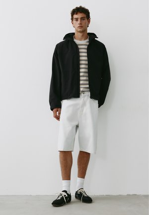 Jeune homme portant une veste noire, une chemise rayée, un short blanc jusqu'aux genoux, des chaussettes blanches et des baskets noires, debout contre un mur blanc.