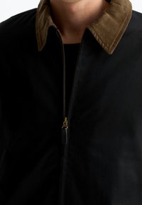 Persoon in een zwarte jas met een bruine corduroy kraag en een gedeeltelijk ingepakte messing rits over een zwart shirt.